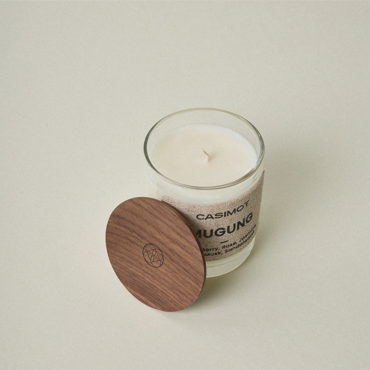 A Casimot Mugung scented candle in a clear glass jar with white wax, shown next to its round wooden lid on a neutral beige background.