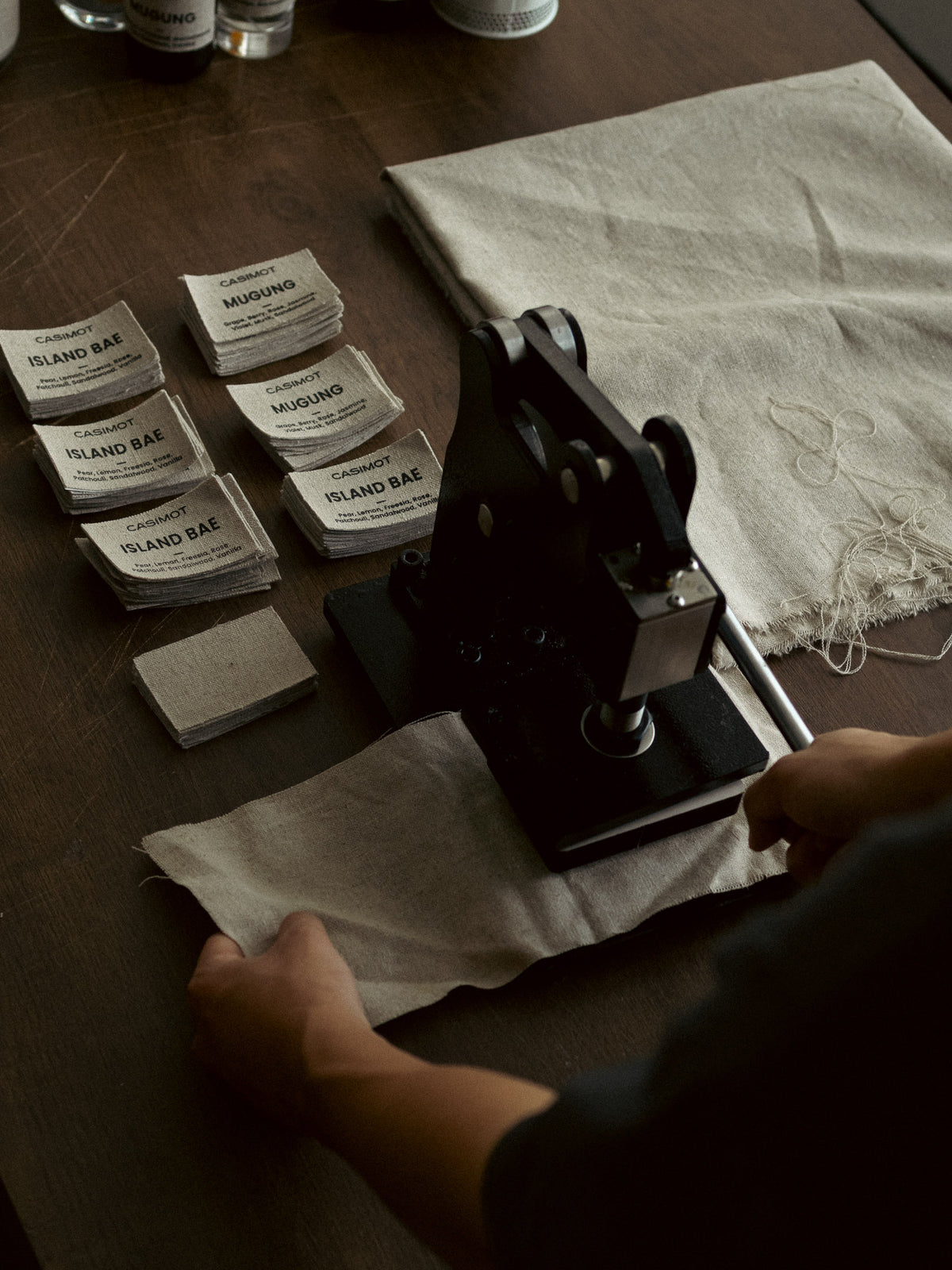 A person's hands guide a piece of linen fabric through a black manual paper cutter on a dark wooden table. Several small printed cards labeled 'CAMPFIRE ISLAND BAE' are stacked nearby, and a folded piece of matching linen sits to the right.