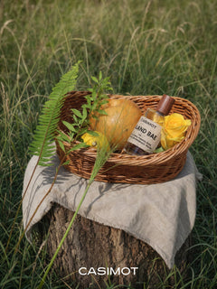 CASIMOT Island Bae home fragrance bottle in a woven basket with Korean pear, yellow roses, and fresh greenery, styled outdoors on a tree stump.