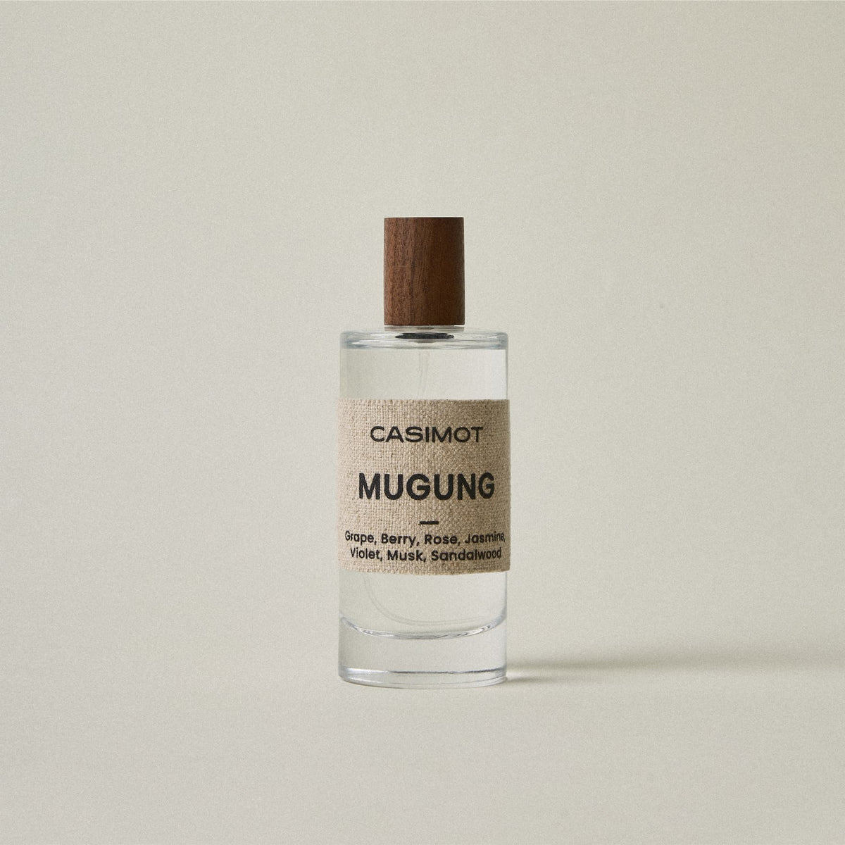 A clear glass bottle of Casimot Mugung perfume with a textured beige label and a dark brown wooden cap, set against a solid light beige background.
