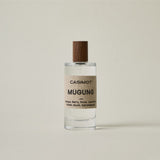 A clear glass bottle of Casimot Mugung perfume with a textured beige label and a dark brown wooden cap, set against a solid light beige background.