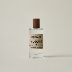 A clear glass bottle of Casimot Mugung perfume with a textured beige label and a dark brown wooden cap, set against a solid light beige background.