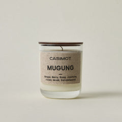 A Casimot Mugung scented candle in a clear glass jar with a natural wood lid and a beige linen label, set against a neutral light beige background.